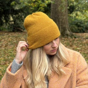 Op de afbeelding: Mosterdgele gebreide beanie. De hoed is gemaakt van een zacht, getextureerd materiaal. De beanie is bij de rand gevouwen en wordt door een persoon aangepast. De achtergrond is een park met bomen.
