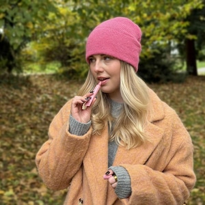 May include: A woman wearing a pink beanie and a peach-colored coat holds a pink lip product. The background features a blurred view of trees and fallen leaves, suggesting an outdoor setting.