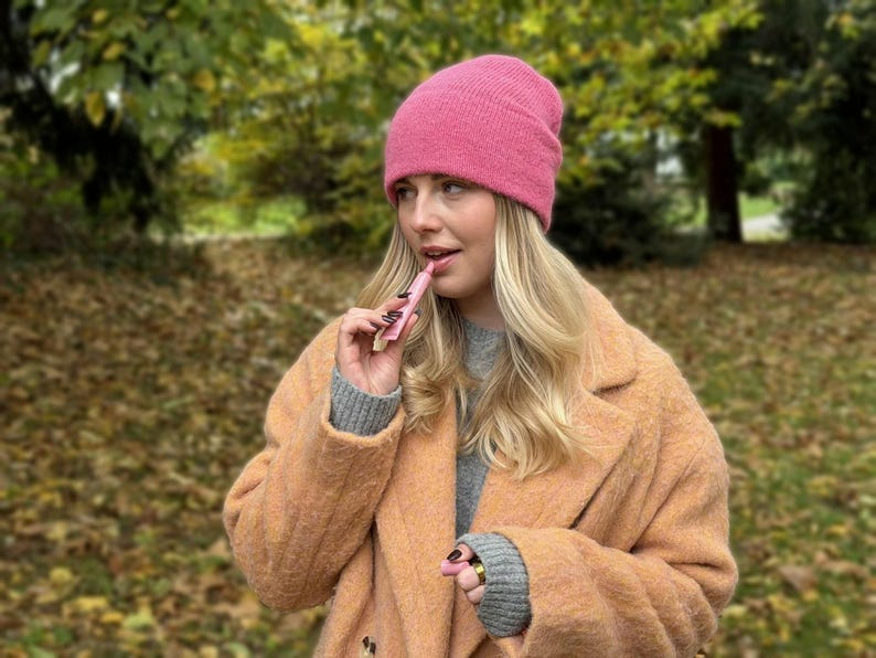 May include: A woman wearing a pink beanie and a peach-colored coat holds a pink lip product. The background features a blurred view of trees and fallen leaves, suggesting an outdoor setting.