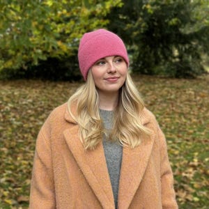 May include: A woman wearing a pink knit beanie, a gray sweater, and a peach-colored coat. The background features fall foliage with green and brown leaves. The beanie is a solid, vibrant pink color.