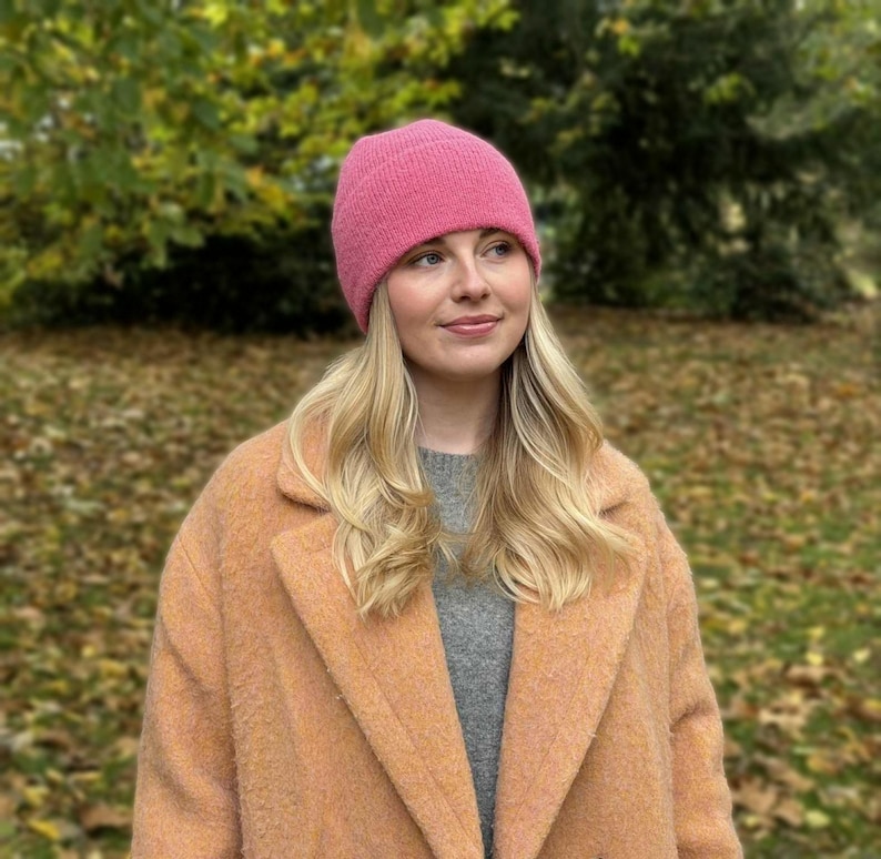 May include: A woman wearing a pink knit beanie, a gray sweater, and a peach-colored coat. The background features fall foliage with green and brown leaves. The beanie is a solid, vibrant pink color.