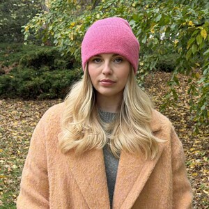 May include: A woman wearing a bright pink knit beanie and a peach-colored coat. The woman has long blonde hair and is looking directly at the camera. The background is a blurred view of a park with green and yellow foliage.