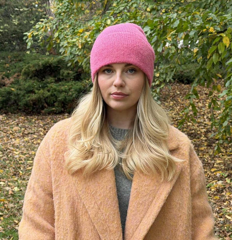 May include: A woman wearing a bright pink knit beanie and a peach-colored coat. The woman has long blonde hair and is looking directly at the camera. The background is a blurred view of a park with green and yellow foliage.
