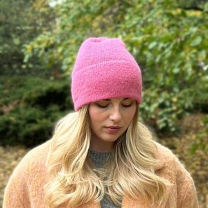 May include: A vibrant pink beanie hat. The hat is made of a soft, textured material. The hat is worn by a person with long blonde hair and a light orange coat. The background is blurred with green foliage.