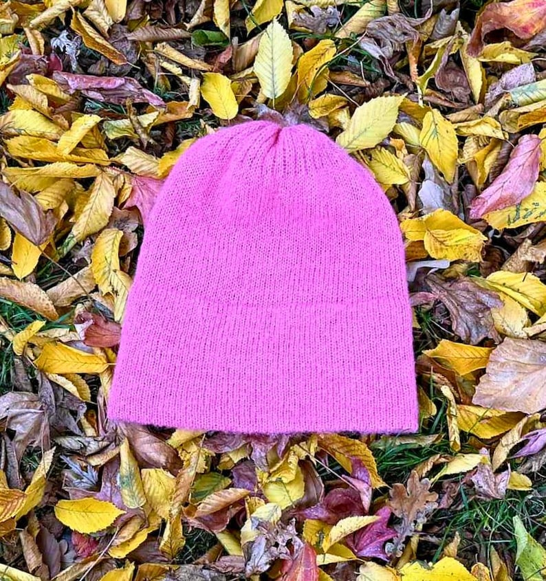 May include: A bright pink knit beanie hat. The hat is made of a soft, textured material and is set against a backdrop of colorful autumn leaves in shades of yellow, brown, and green. The hat is a solid color and has a simple, classic design.