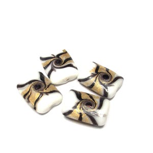 May include: Four square-shaped beads with a white base, adorned with gold and black spiral and stripe designs. The beads are scattered against a white background, showcasing their unique patterns and textures.