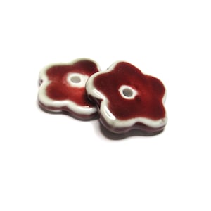 May include: Two ceramic flower-shaped beads. The beads are a deep red color with a white border and a hole in the center. The beads are on a white background.