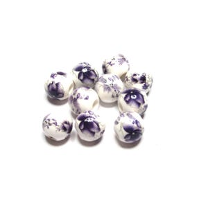 May include: A collection of white ceramic beads with a floral pattern in shades of purple. The beads are round and have a slightly irregular shape, with the floral design covering the surface.
