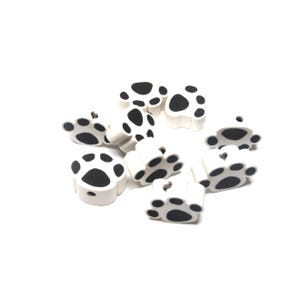 May include: A group of white ceramic beads with black paw print designs. The beads have a hole in the center for stringing.