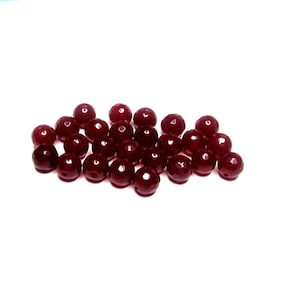 May include: A collection of faceted, round beads in a deep burgundy color. The beads are scattered against a white background, showcasing their polished surfaces and spherical shape. These beads are suitable for jewelry making.