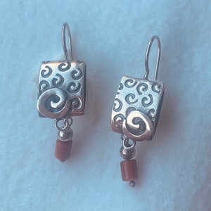 May include: Silver dangle earrings with a square-shaped charm featuring a swirl design and a brown stone bead.