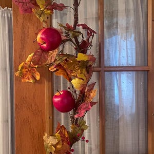 May include: A decorative autumn garland featuring artificial red apples, small red berries, and a mix of yellow, orange, and deep red maple-style leaves. The garland is constructed on a brown, branch-like wire frame with a small white tag attached.