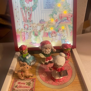 May include: A Christmas-themed display box with a red frame, featuring a painted winter scene with a Christmas tree and toys. The box contains figurines of Santa Claus, Mrs. Claus, a reindeer, and other characters. The words "Merry Christmas" are printed on a decorative sign.