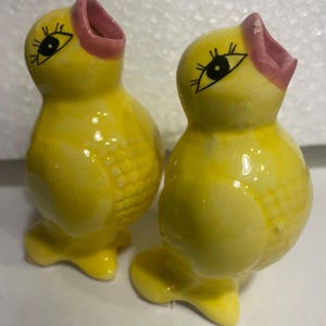 May include: A pair of yellow ceramic chick figurines with pink open beaks and black painted eyes with long eyelashes. Each bird has a textured wing detail on the side and stands on a small base. The glossy finish reflects light on the rounded bodies.