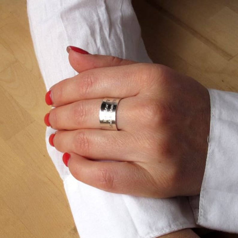 May include: A silver band ring with an inscription on the side. The ring is worn on the middle finger of a hand with red nail polish.