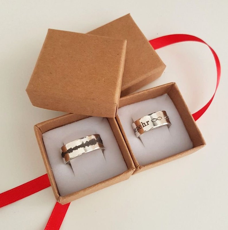 May include: Two small brown boxes with white interiors, each containing a silver ring. One ring has a black sound wave design, and the other has the word "forever" and an infinity symbol engraved on it.