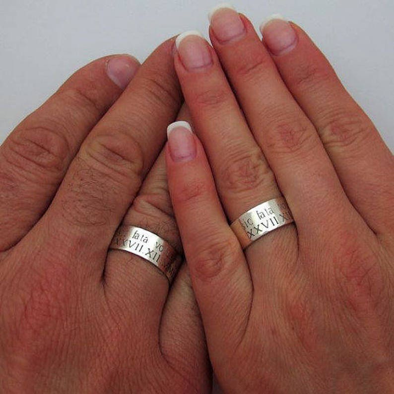 May include: Two silver wedding bands with Roman numeral engravings. The bands are worn on the ring fingers of two hands.