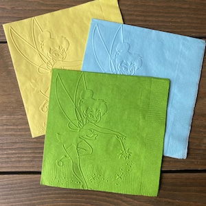 May include: Three paper napkins with a green, yellow, and blue background. Each napkin features an embossed image of Tinkerbell, a fairy with wings, holding a wand and a star.