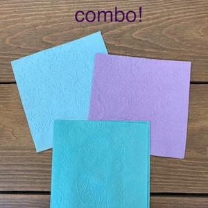 May include: Three paper napkins with a Frozen theme. The napkins are blue, purple, and teal. The blue napkin has a snowflake pattern, the purple napkin has a snowflake pattern, and the teal napkin has an image of Elsa from Frozen.