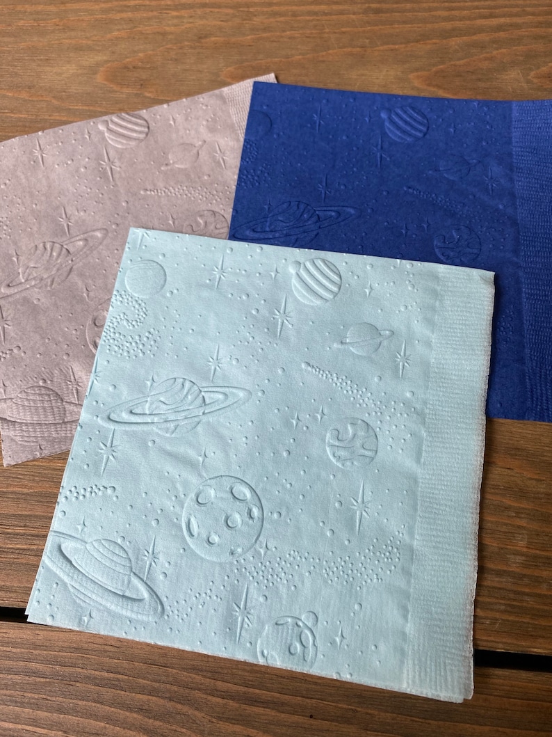 May include: Three paper napkins with an embossed space theme design. The napkins are in shades of blue, gray, and light blue. The design features planets, stars, and moons.