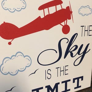 May include: Canvas art with a red biplane, blue clouds, and the navy blue text "The Sky is the Limit". The artwork has a white background and a simple design. The piece is suitable for a child's room.