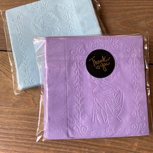May include: Two packages of blue and purple paper napkins with an embossed design. The purple napkins have a gold sticker that says "Thank You".