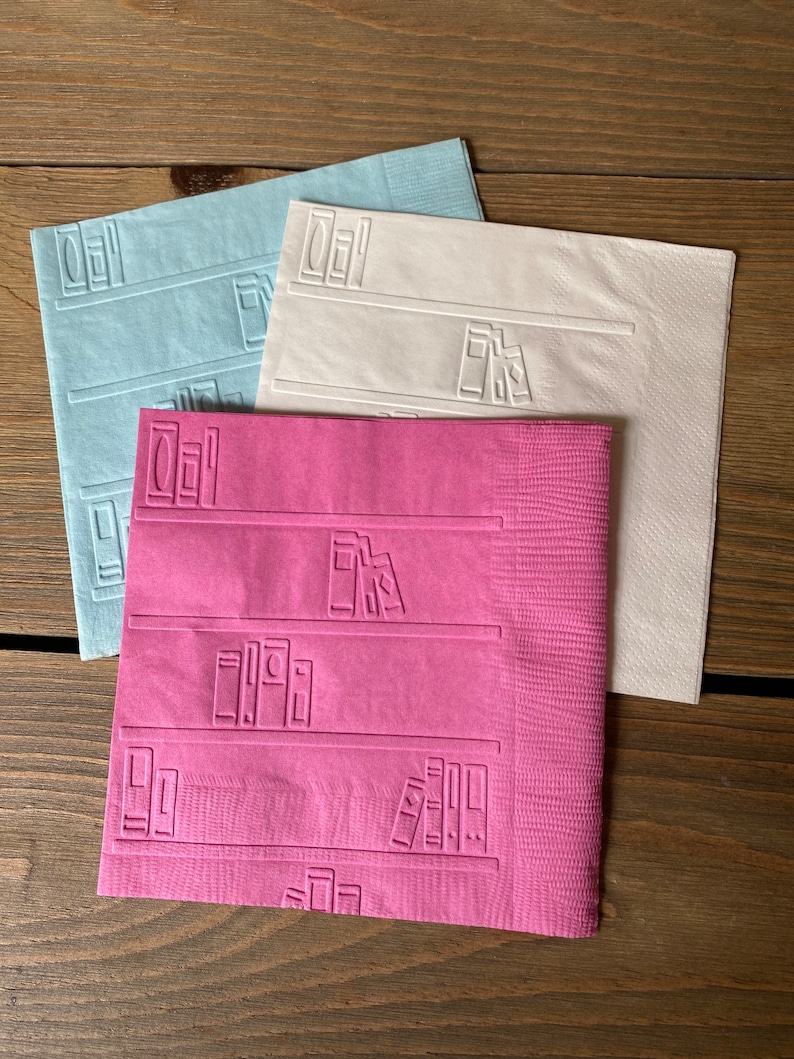 May include: Three paper napkins with an embossed design of books on a bookshelf. The napkins are in blue, white, and pink.