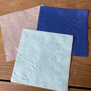 May include: Three paper napkins with a space theme. The napkins are blue, light blue, and gray. Each napkin has a pattern of planets, stars, and comets.
