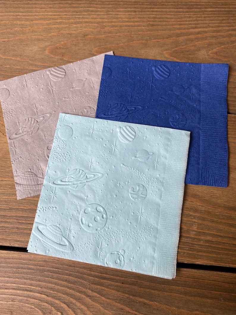 May include: Three paper napkins with a space theme. The napkins are blue, light blue, and gray. Each napkin has a pattern of planets, stars, and comets.