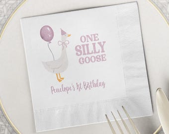 May include: White paper napkin featuring a cartoon goose with a party hat and balloon. The text reads "ONE SILLY GOOSE" and "Penelope's 1st Birthday" in pink. The napkin is placed on a white plate with cutlery.