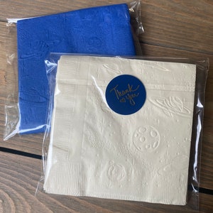 May include: Two packages of white paper napkins with a blue embossed design of planets and stars. One package has a blue circle sticker with the text "Thank you" in gold lettering.