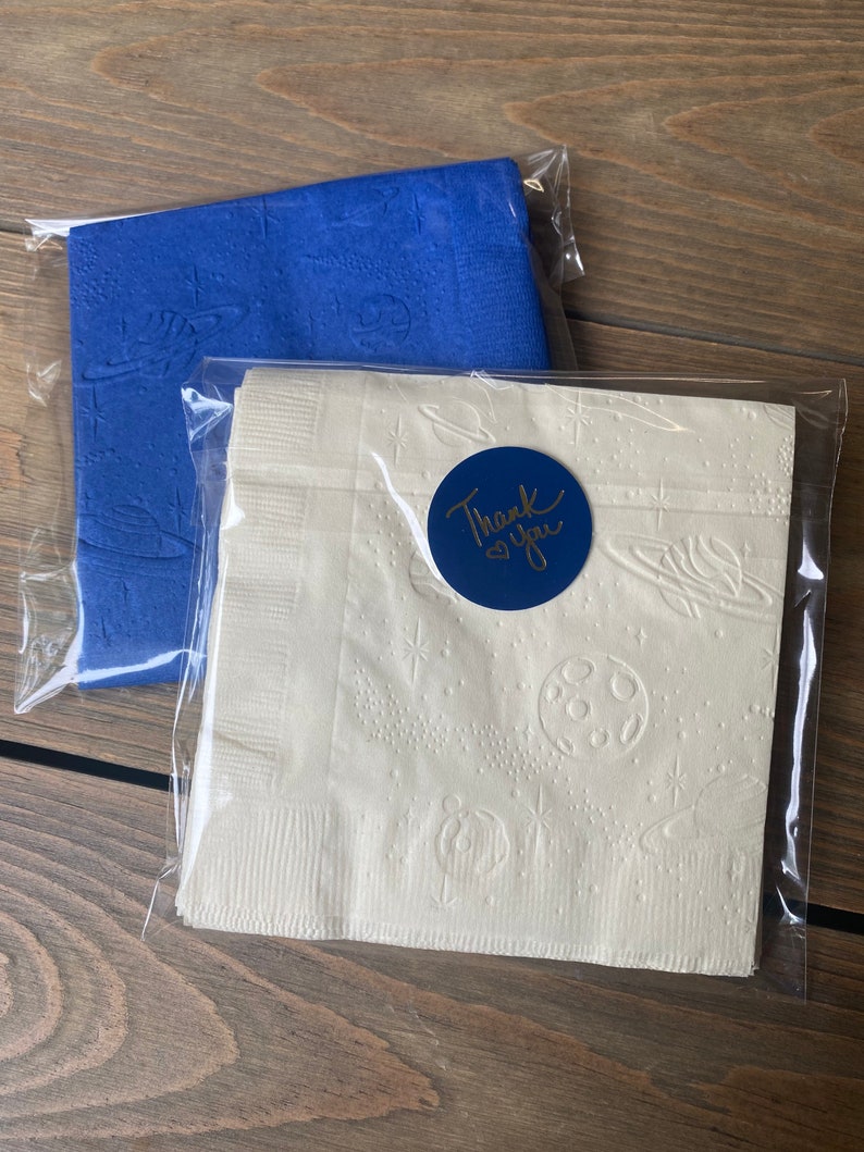 May include: Two packages of white paper napkins with a blue embossed design of planets and stars. One package has a blue circle sticker with the text "Thank you" in gold lettering.