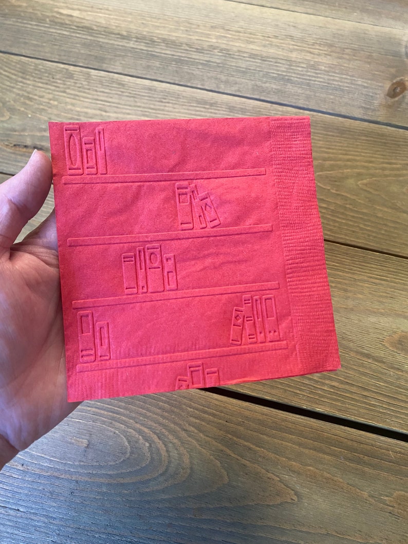 May include: A red paper napkin with a bookshelf design embossed on it. The bookshelf has four shelves with books on each shelf.