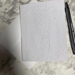 May include: A white embossed greeting card featuring a portrait of a princess within a floral frame, accompanied by a matching envelope. A silver pen rests on the surface. The card's design is detailed and elegant, suitable for special occasions.