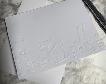 May include: A white card with embossed woodland animal designs, including a bear, fox, and rabbit, rests on top of a white envelope. A silver pen is positioned to the right. The card and envelope are on a marble-patterned surface.