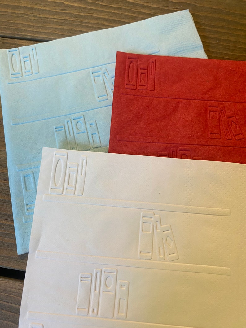 May include: Three decorative paper placemats with an embossed design of books on a bookshelf. The placemats are in blue, red, and white.