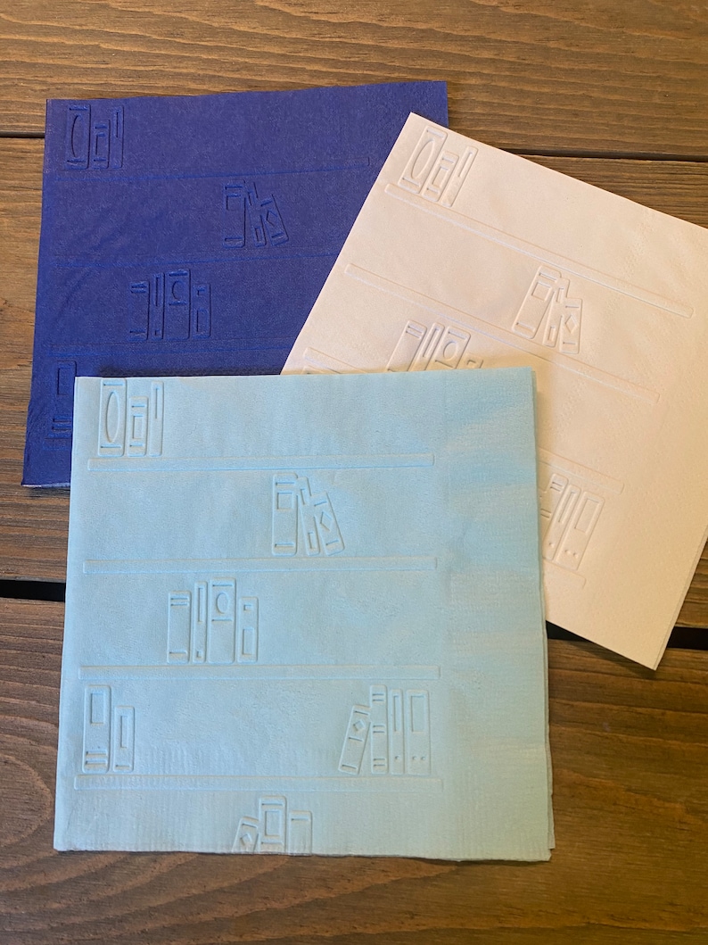 May include: Three paper napkins with a bookshelf design. The napkins are blue, white, and light blue. Each napkin has a repeating pattern of books on a shelf.