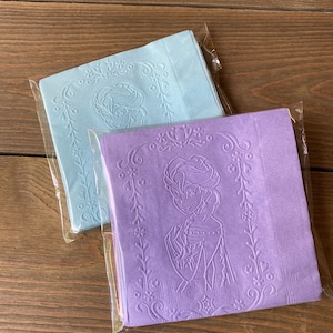 May include: Two packages of paper napkins with an embossed design of a cartoon character. One package is light blue and the other is purple.