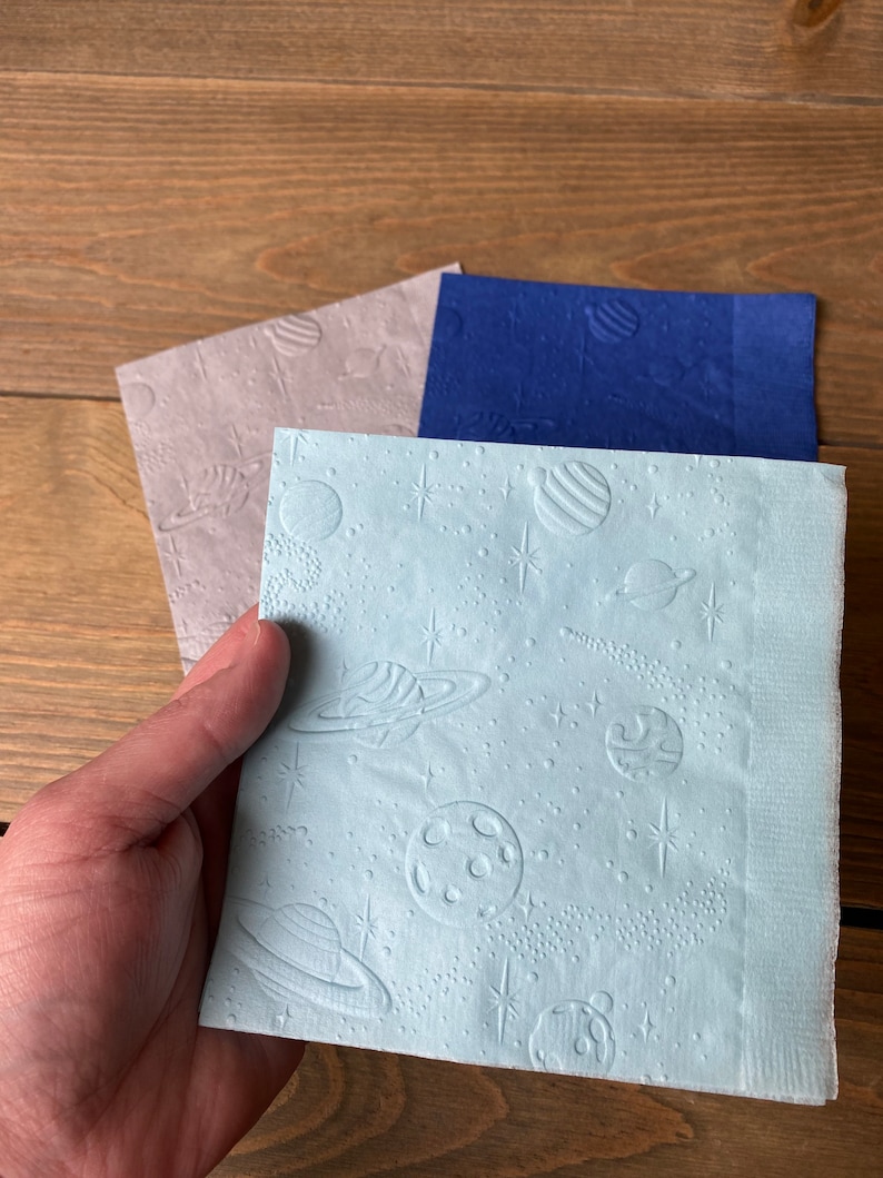 May include: Three paper napkins with a space theme. The napkins are light blue, dark blue, and light brown. Each napkin has a different design of planets, stars, and moons.