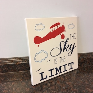 May include: A canvas art piece with a cream background features a red biplane, blue clouds, and the phrase "The Sky is the Limit" in navy blue. The artwork is displayed on a dark brown surface.