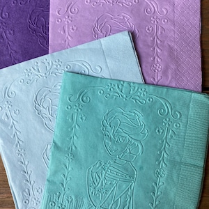 May include: Four paper napkins with an embossed design. The napkins are in shades of purple, light blue, and green. The green napkin features an embossed image of a woman with long hair and a crown.