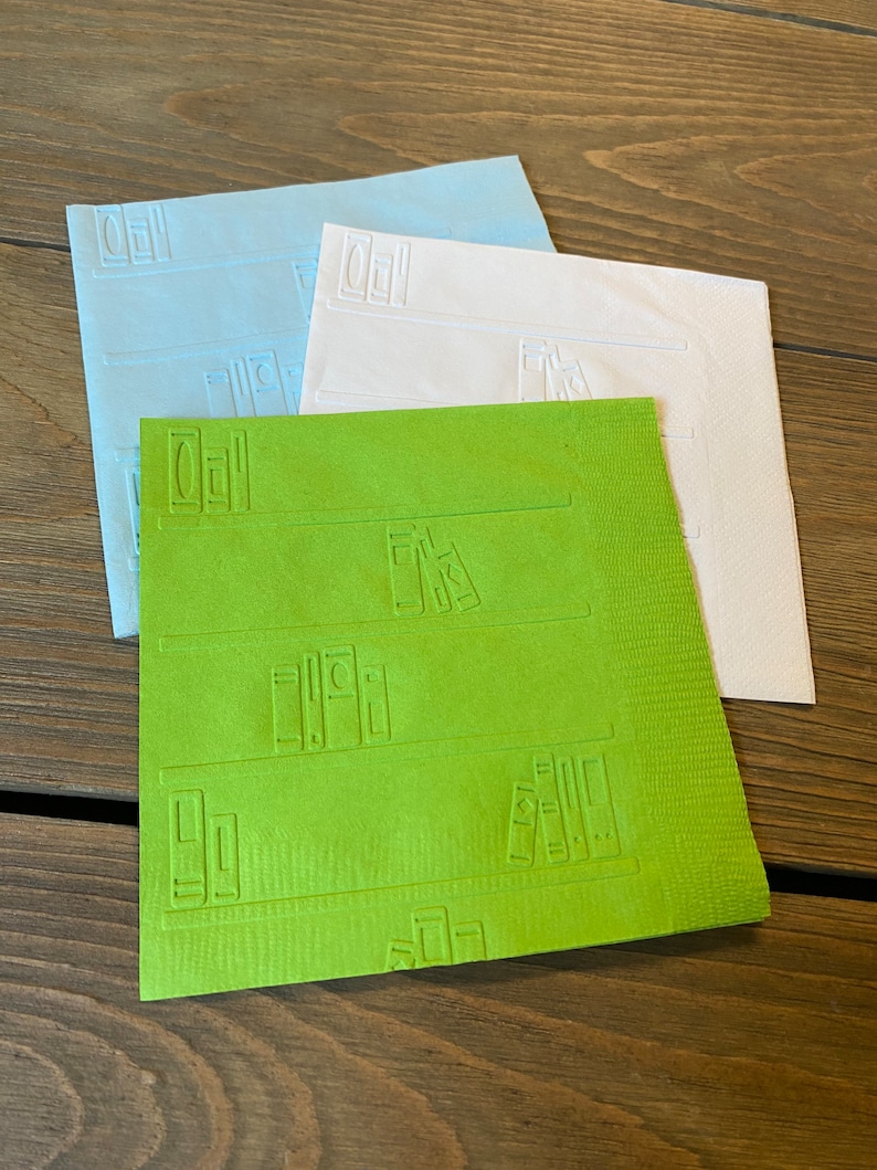 May include: Three paper napkins with an embossed design of books on a bookshelf. The napkins are in shades of blue, white, and green.