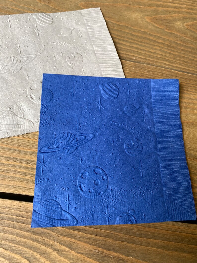 May include: Two paper napkins with an embossed space theme. One napkin is gray with a white embossed design, and the other is blue with a white embossed design.