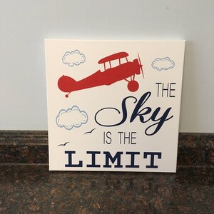 May include: A square, off-white canvas art piece featuring a red aeroplane silhouette, blue clouds, and the phrase "The Sky is the Limit" in navy blue. Two small birds are also depicted.
