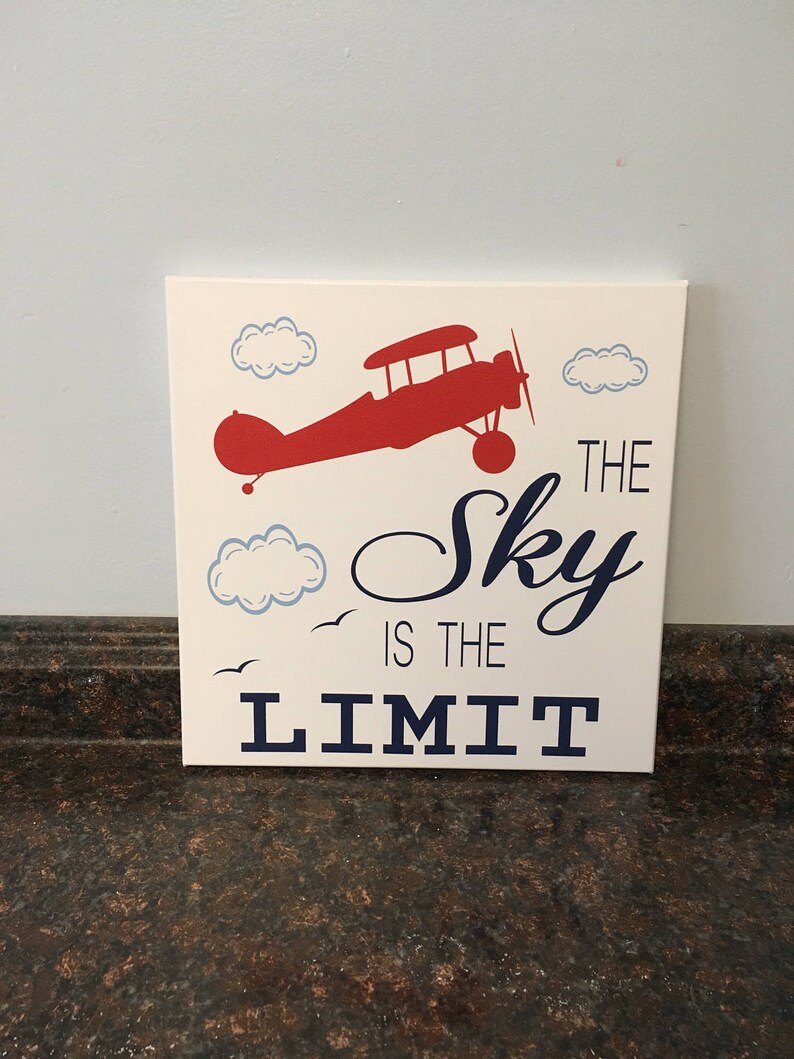 May include: A square, off-white canvas art piece featuring a red aeroplane silhouette, blue clouds, and the phrase "The Sky is the Limit" in navy blue. Two small birds are also depicted.