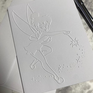 May include: A white card with an embossed image of Tinkerbell, a fairy with wings, holding a wand and sprinkling pixie dust.