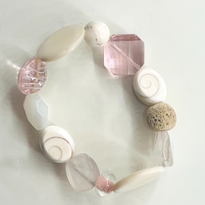 Könnte beinhalten: Ein handgefertigtes Armband mit einer Mischung aus weißen, rosa und beigen Perlen. Das Armband hat verschiedene Formen, darunter oval, rund und geometrisch, sowie einige muschelförmige Perlen. Der Umfang beträgt ca. 18 cm.