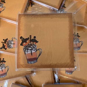 May include: A stack of brown paper notepads with a whimsical illustration of a coffee drink topped with whipped cream, chocolate sauce, and candy eyeballs. The notepads are individually wrapped in clear plastic.