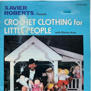 May include: A book cover featuring a man in a black hat and suit holding a doll. The book title is "Crochet Clothing for Little People" with the subtitle "with Shirley Now". The cover also features a dollhouse and several dolls.