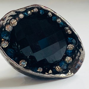 Peut inclure: Une bague en argent avec une grosse pierre précieuse noire entourée d'un anneau de strass bleus et clairs.
